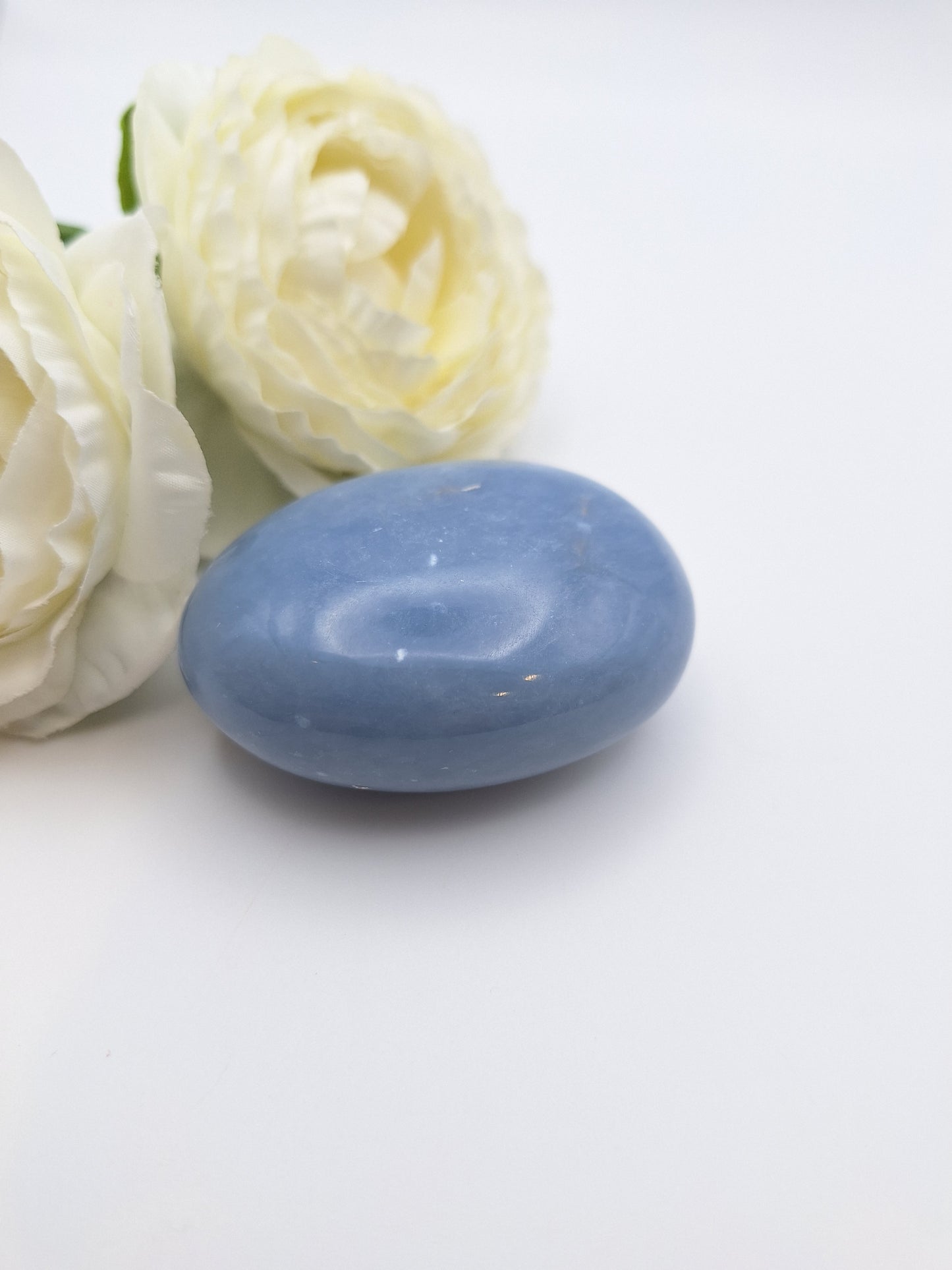 A single Angelite palm stone next to a delicate white flower, highlighting its soft blue color and serene energy.