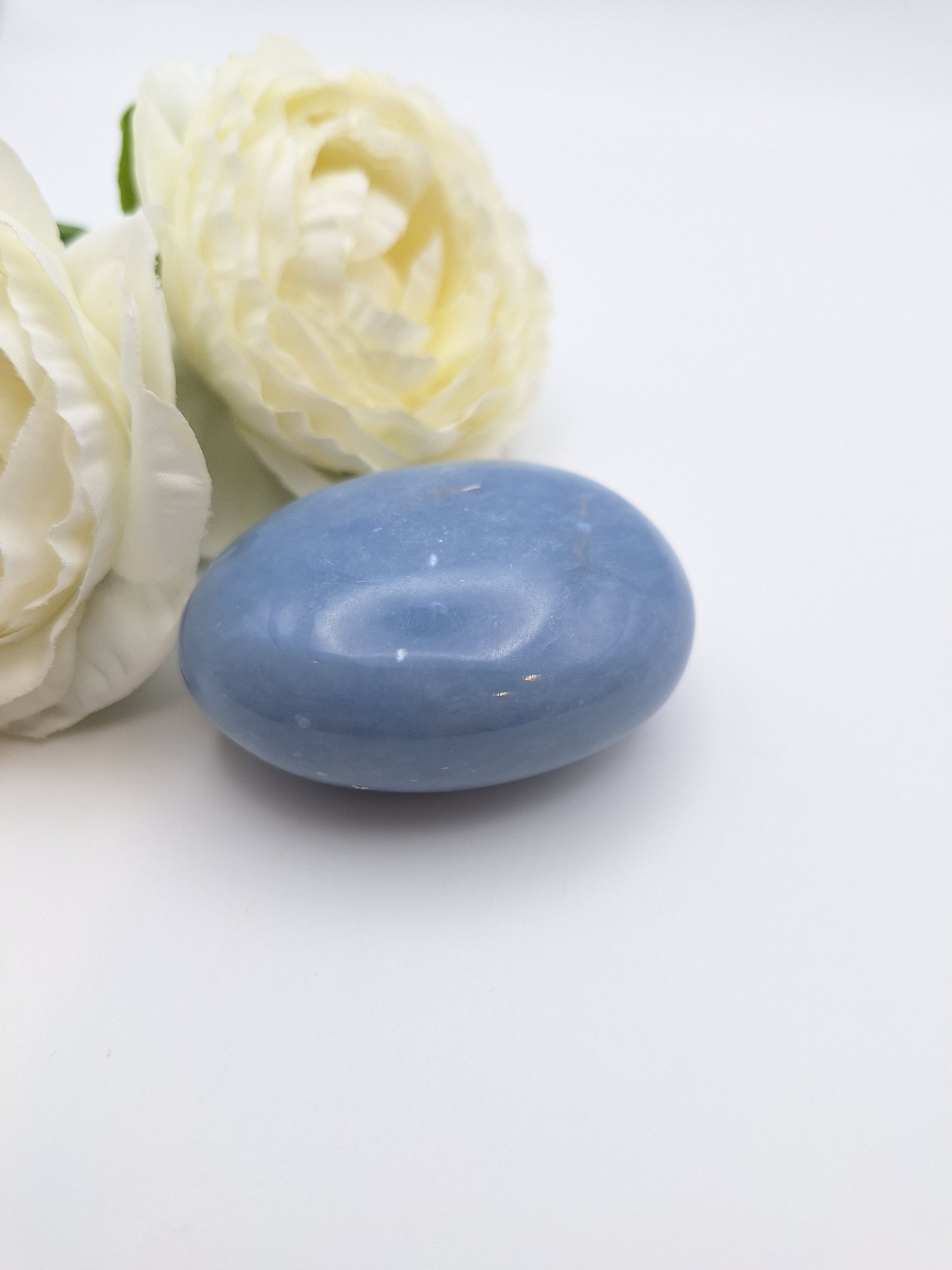 A single Angelite palm stone next to a delicate white flower, highlighting its soft blue color and serene energy.