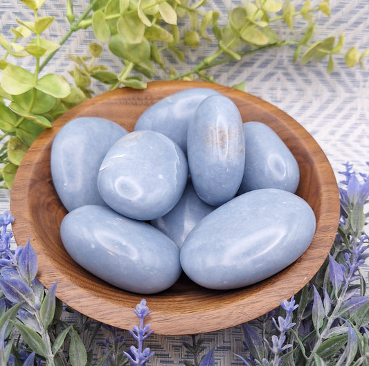 A wooden bowl filled with polished Angelite palm stones from Peru, displaying their soft powder-blue color and smooth finish.