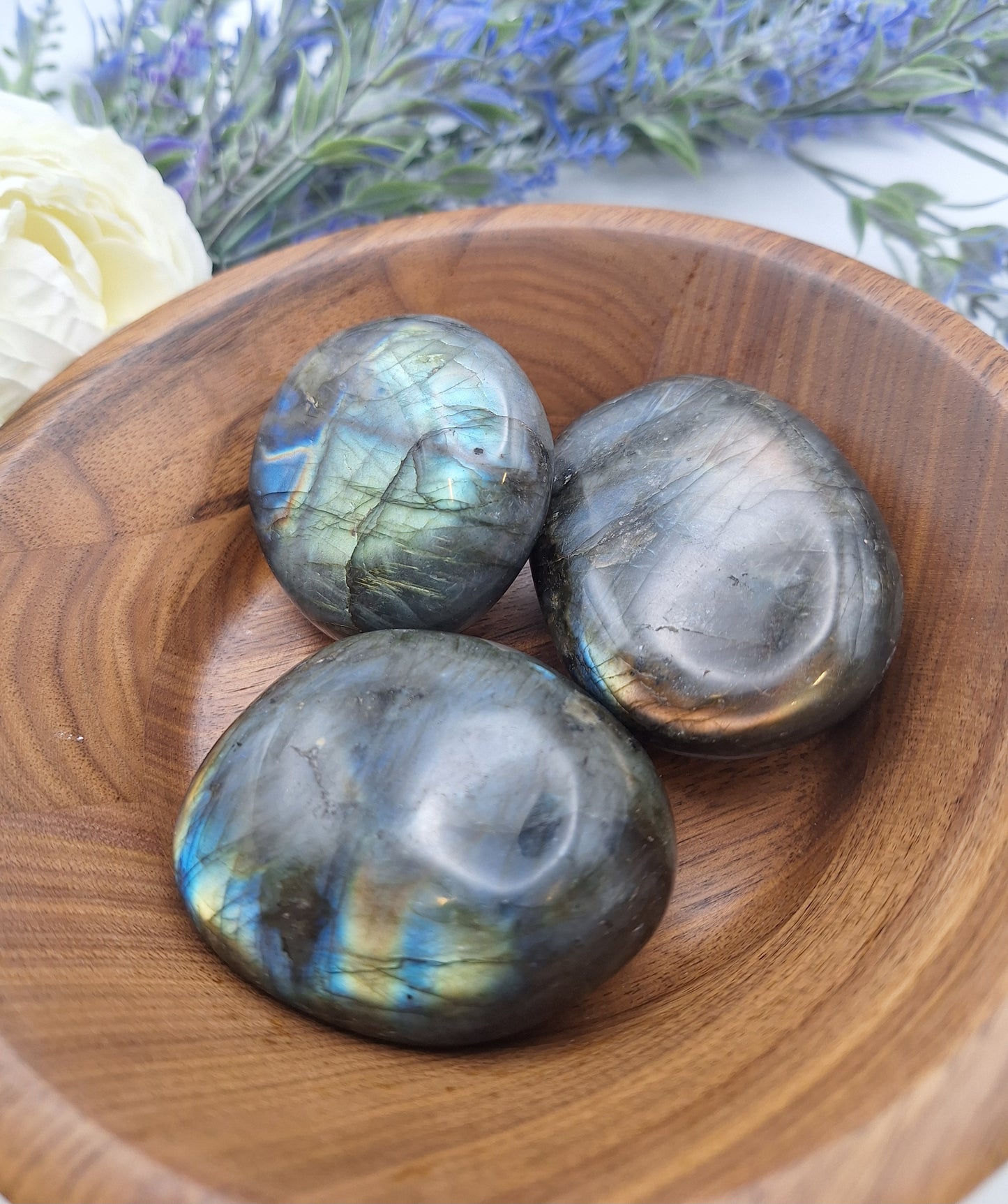 A wooden bowl filled with three polished Labradorite palm stones, showcasing their deep gray base tones with flashes of color.