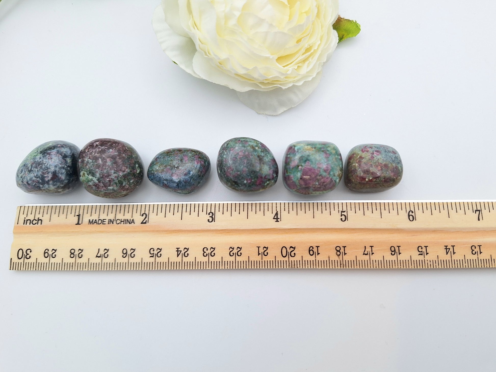 A row of Ruby Kyanite tumbled stones placed along a ruler for size reference, showcasing their smooth, polished surface and natural color variations.